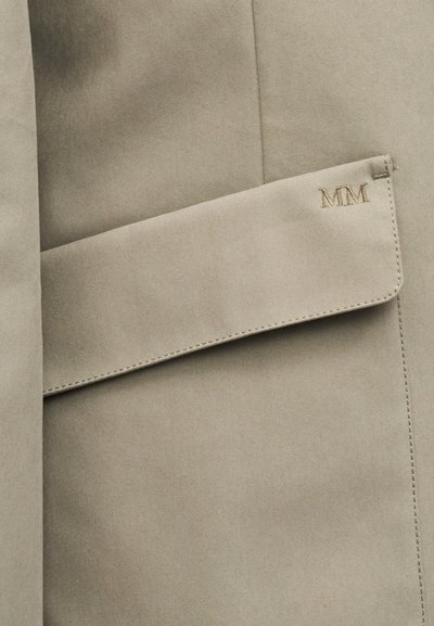 Κοντινό πλάνο της τσέπης ενός μπεζ παλτού τύπου trench, με ραμμένο καπάκι και κεντημένα αρχικά "MM" στην ανώτερη γωνία.
