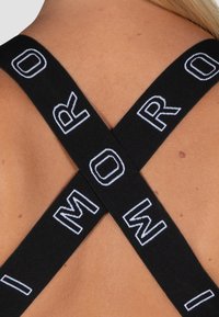 Tiras elásticas negras forman una forma de X en la parte posterior, con letras blancas tejidas en el material. La piel suave es visible alrededor de las tiras.