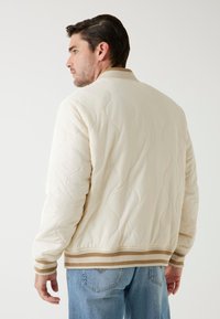 Překrytá slonovinová bunda bomber s hnědobílými proužky na lemu a manžetách. Má hladkou texturu a klasický design límce.