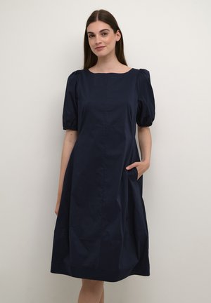 Robe en coton bleu marine avec un décolleté carré, des manches courtes bouffantes et des poches de chaque côté. Texture lisse, silhouette en A.