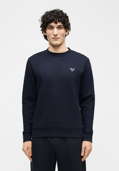 Άνδρας με σγουρά μαλλιά που φοράει ένα απλό σκούρο μπλε φούτερ με ένα μικρό σύμβολο καρδιάς στο αριστερό στήθος, στέκεται μπροστά σε λευκό φόντο.