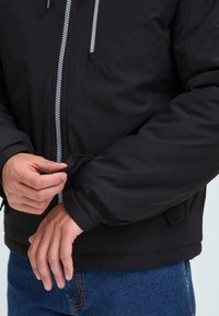 Hands adjusting wrist strap of black jacket with front zipper, worn over blue jeans.
