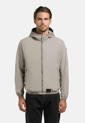 Mężczyzna w jasnoszarej kurtce z kapturem na suwak i czarnych spodniach, stoi na jednolitym białym tle, zwrócony przodem z neutralnym wyrazem twarzy.