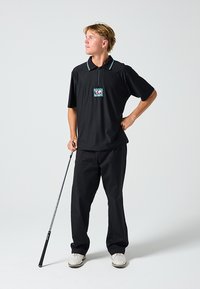 Μαύρο κοντομάνικο πόλο με φερμουάρ στον γιακά και πολύχρωμο γραφιστικό λογότυπο, σε συνδυασμό με μαύρο παντελόνι και λευκά αθλητικά παπούτσια, κρατώντας ένα μπαστούνι.