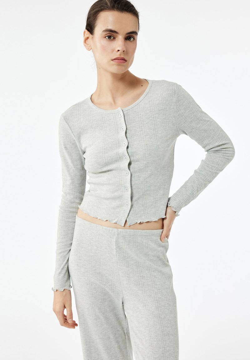 Grå ribbet cardigan med en cropped pasform, rund halsudskæring og knaplukning fortil. Har lange ærmer og bølgede kantdetaljer.