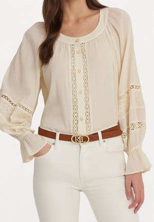 Mujer que lleva una blusa color crema con inserciones de encaje, mangas largas abullonadas y botones delanteros, combinada con pantalones blancos y un cinturón marrón con hebilla dorada.