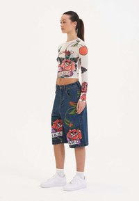 Langarmige doorschijnende top met bloemenprint en cropped ontwerp, gecombineerd met denimshorts met grote bloemenillustraties en tekstaccenten.