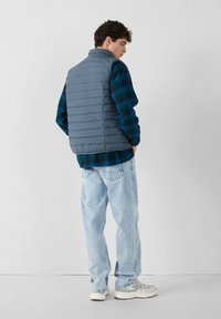 Chaleco de plumas gris con un cuello alto sobre una camisa de franela a cuadros azul y verde. Unos jeans azul claros y zapatillas grises completan el atuendo.
