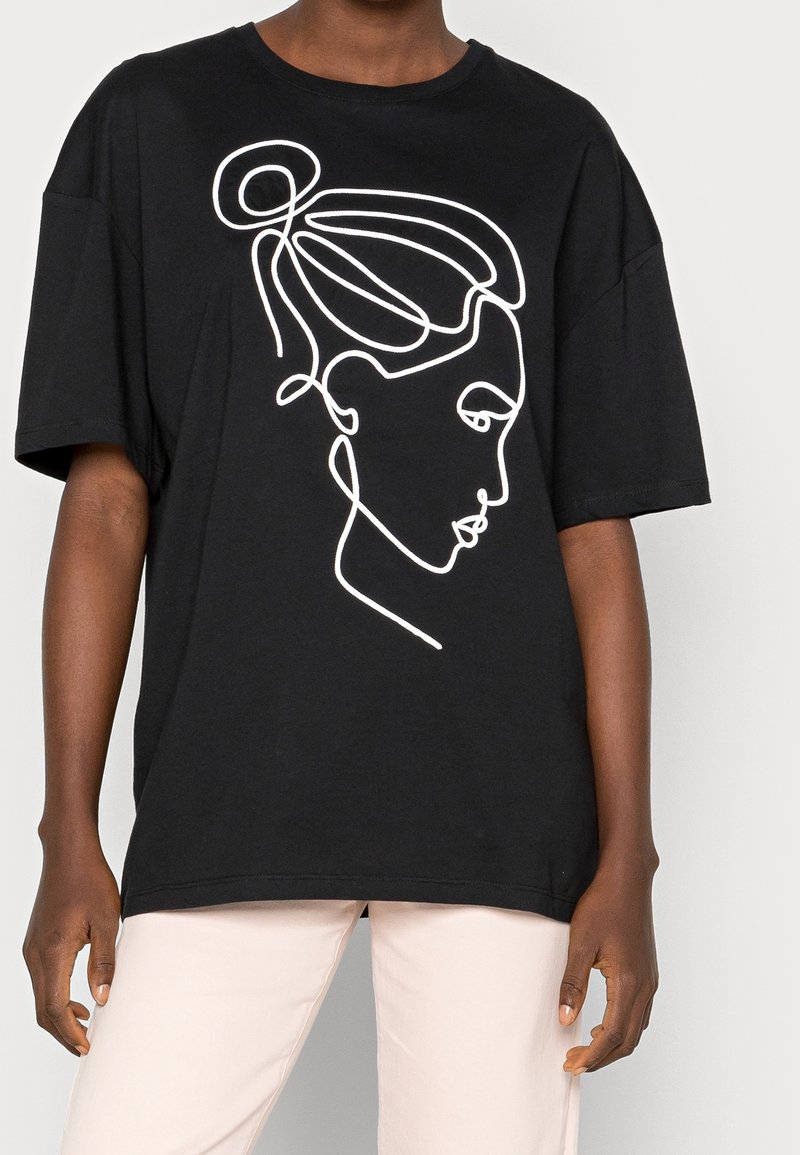 Zwart oversized T-shirt met een witte doorlopende lijntekening van het profiel van een vrouw met haar in een knot, gedragen met lichtbeige broek.