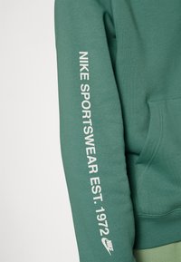 Grön sweatshirtärm med vit text "NIKE SPORTSWEAR EST. 1972" och Nike-logotyp nära handleden, som visar en del av en framficka.