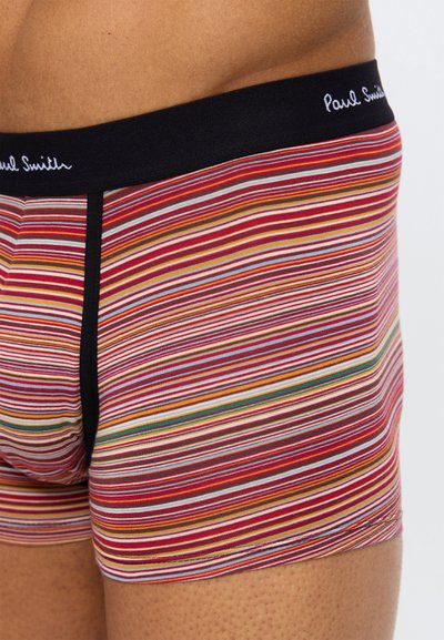 Gros plan sur une personne portant un boxer rayé multicolore avec une ceinture noire portant l'inscription "Paul Smith".