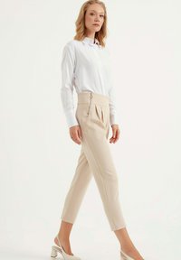 Lichtbeige high-waisted broeken met plooien en een ritsdetail, gecombineerd met een witte langslavige overhemd met knopen en lichtgekleurde haksandalen.