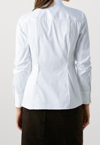 Biała dopasowana koszula z długim rękawem, detal w postaci karczku z tyłu oraz gładka tekstura; zestawiona z ciemną spódnicą.