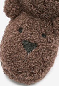 Chausson en peluche brun avec une surface texturée et douce. Présente un design de visage d'ours, incluant des caractéristiques faciales brodées et des oreilles arrondies.