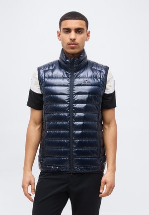 Man draagt een glanzend marineblauw doorgestikt mouwloos vest over een zwart-wit shirt met korte mouwen, staand tegen een effen lichte achtergrond.