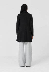 Zwarte op maat gemaakte jas met een ingesneden kraag, enkele split aan de achterkant, gedragen over brede grijze broek. Heeft knoopaccenten op de manchetten.