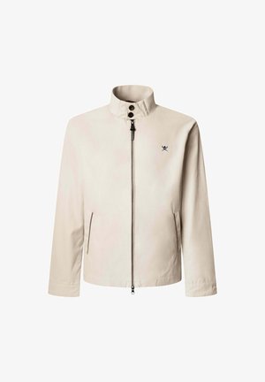 Beige jas met een staande kraag, een ritssluiting aan de voorkant en twee zijzakken. Heeft een klein zwart embleem op de borst. Gladde stofstructuur.