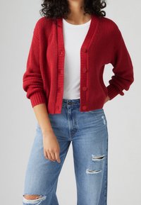 Punainen ribbireunainen villatakki, jossa on V-päänti, nappikiinnitys ja pitkät hihat. Se on yhdistetty valkoiseen toppiin ja revenneisiin sinisiin farkkuihin.