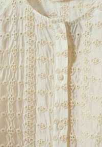 Valkoinen brodeerattu pusero, jossa on monimutkaisia pitsikuvioita, pieniä nappeja ja pehmeä, teksturoitu kangas edessä.