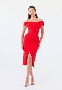 Robe rouge décolletée avec bodice froncé, texture drapée, fente latérale et silhouette ajustée. Associée à des talons nus à lanières.