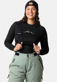 Schwarzes Langarmoberteil mit Berggrafik, kombiniert mit hellgrünen Überhosen mit Trägern, Knöpfen und seitlichen Reißverschlusstaschen.