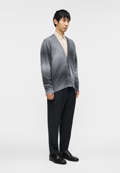 Γκρι πλεκτή ζακέτα με V-λαιμόκοψη, κουμπιά και μακριά μανίκια, φορεμένη πάνω από ανοιχτόχρωμο πουκάμισο· συνδυασμένη με σκούρο παντελόνι με ρίγες και μαύρα παπούτσια.