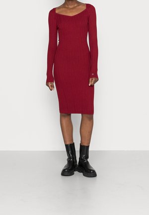 Punainen neulemekko, jossa on pyöreä pääntie ja pitkät hihat. Mekossa on ribbineulepinta ja se on kapeamallinen. Asusteena mustat nilkkasukat.