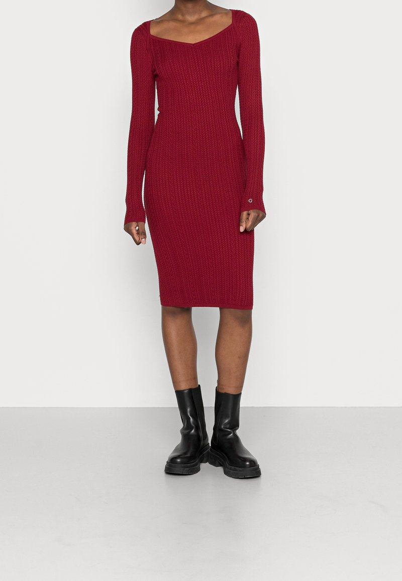 Punainen neulemekko, jossa on pyöreä pääntie ja pitkät hihat. Mekossa on ribbineulepinta ja se on kapeamallinen. Asusteena mustat nilkkasukat.