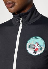 Crna jakna na zip s visokim ovratnikom, koja ima bijeli zip i vezeni okrugli patch s prikazom skijaša u sjedećem položaju.