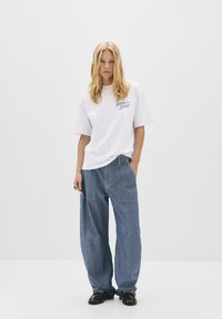 T-shirt blanche avec texte bleu, associé à un pantalon large rayé bleu. La tenue présente une coupe décontractée et un design casual avec des poches.