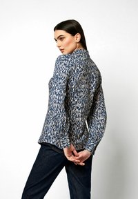Langärmlige Bluse mit einem blauen und cremefarbenen abstrakten Muster, einem kragenlosen Ausschnitt und gerafften Bündchen. Getragen mit dunklen Denim-Hosen.