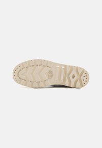 Suela de una zapatilla en goma de color beige claro con patrones de textura en la banda de rodadura y ranuras de soporte. Presenta una marca en la entresuela, diseño limpio.