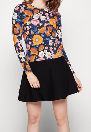 Frau trägt ein langärmeliges, geblümtes Oberteil mit orangefarbenen, weißen, rosa und blauen Blumen kombiniert mit einem schwarzen A-Linien-Rock.