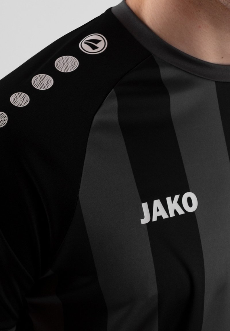 Maglietta sportiva nera con strisce verticali grigie. Presenta un colletto rotondo, accenti strutturati sulle spalle e il logo bianco "JAKO" sul davanti.