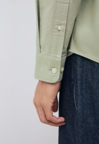 Camicia verde chiaro a maniche lunghe con dettaglio del polsino abbottonato, abbinata a denim scuro. Il tessuto appare liscio e strutturato.