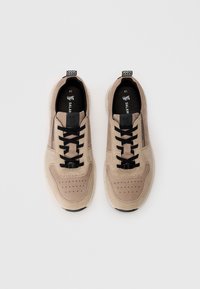 Beige Wildleder-Sneaker mit schwarzen Schnürsenkeln und Akzenten, durchlöchertem Zehenbereich und strukturiertem Außensohlen. Mit einem Logo-Patch in der Nähe des oberen Teils.