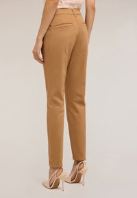 Pantaloni in misto cotone marrone con una vestibilità dritta, gambe affusolate, due tasche posteriori e zip alla caviglia. Indossati con scarpe col tacco nude.