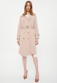 Beżowy płaszcz trench z lekkiego materiału, z podwójnym zapięciem, złotymi guzikami, talią z paskiem oraz klasycznym kołnierzem.