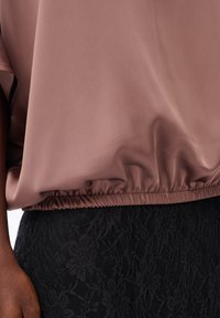 Bruine losse top met een gathered zoom, gedragen over een zwarte bloemen kanten rok, met zichtbare arm en hand van de persoon die de kleding draagt.