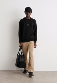 Μαύρη φούτερ μπλούζα με τετράγωνο λογότυπο, μπεζ παντελόνι και μαύρα αθλητικά παπούτσια. Το μοντέλο κρατάει μια μαύρη τσάντα με δύο χερούλια. Απλός, άνετος σχεδιασμός.