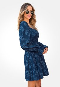 Vestido azul marino con patrón floral y mangas largas abullonadas; tejido texturizado; dobladillo de falda en capas. Combinado con gafas de sol redondas, cabello suelto.