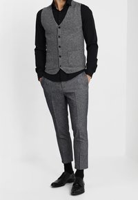 Chaleco gris texturizado con botones negros sobre una camisa negra, combinado con pantalones grises con patrón y zapatos de cuero negros.