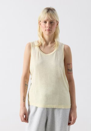 Blond osoba v bezrukávovom bledožltom topu a svetlosivých nohaviciach, stojaca pred jednoliatym bielym pozadím s viditeľnými tetovaniami na rukách.