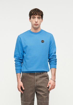 Junger Mann trägt ein blaues Sweatshirt mit kleinem rundem Logo auf der Brust und braune Hose, steht vor einem schlichten hellen Hintergrund.