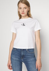 Balti medvilnės trumpomis rankovėmis marškinėliai su juodu logotipu, kuriame centre yra užrašas "CK", ir trumpu apatini kraštu, derinami su mėlynomis džinsomis.