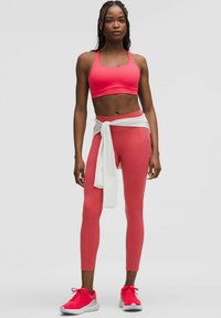 Sujeción deportiva rosa ajustada y leggins de cintura alta, ambos hechos de tejido elástico, con una chaqueta blanca atada a la cintura y zapatillas deportivas rojas.