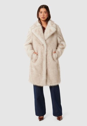 Femme portant un long manteau en fausse fourrure beige clair avec un large col, un pantalon bleu foncé à jambes larges et des chaussures pointues marron, debout les mains dans les poches.
