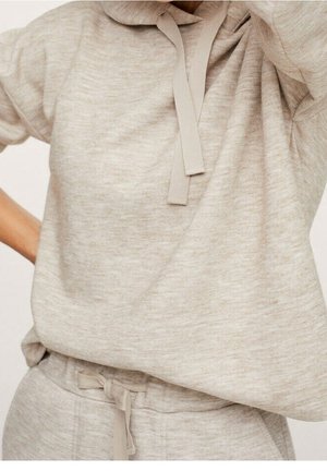 Personne portant un pull en maille beige avec un col court et un pantalon assorti à cordon, main sur la hanche, sur un fond uni.