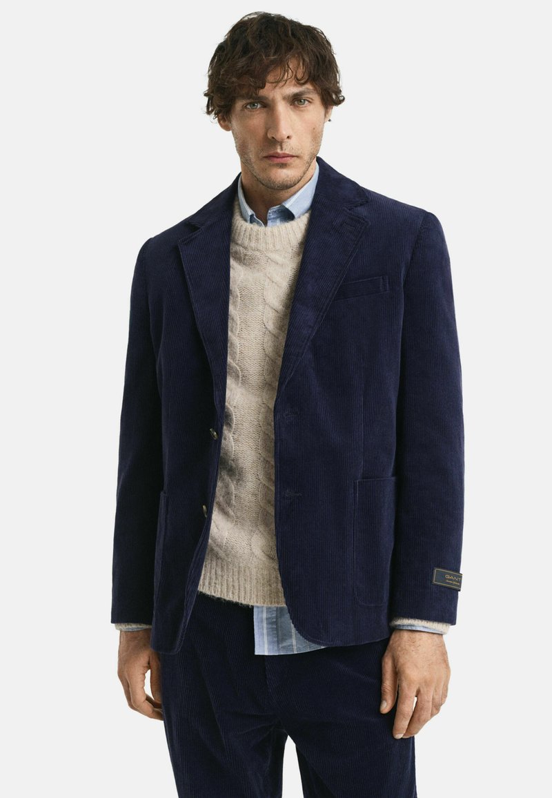 Marineblauer Cordblazer mit zwei Hosentaschen und goldfarbenen Beschlägen, kombiniert mit einem cremefarbenen Grobstrickpullover und einem hellblauen Hemd.