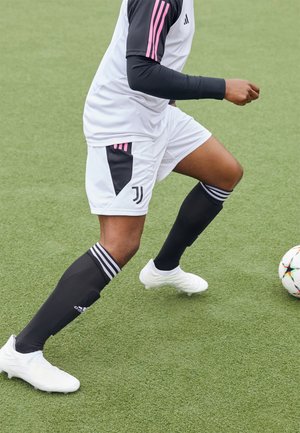 Joueur de football portant un uniforme blanc et noir avec des rayures roses contrôlant un ballon sur un terrain en gazon synthétique vert.